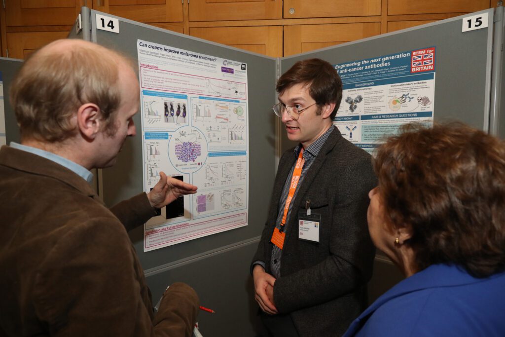Former PhD student Charles Earnshaw standing in front of his research poster and discussing his work with a male and female MPs at the STEM for Britain event in parliament.